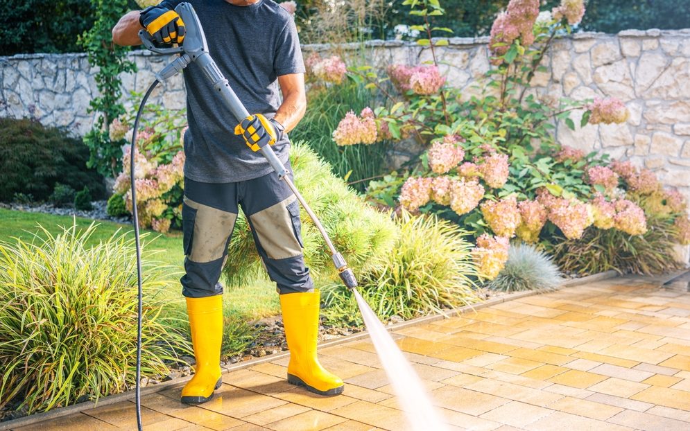 Apersonwearingyellowbootsandglovesoperatesapressure | h3 outdoor design & construction | h3 outdoor design & construction a Person Wearing Yellow Boots and Gloves Operates a Pressure Washer While Cleaning a Paved Patio in a Lush Garden