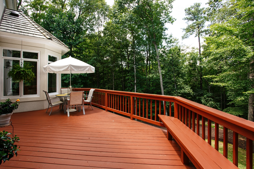 Largewoodendeckfeaturinganumbrella-coveredoutdoordiningsetsurrounded | h3 outdoor design & construction | h3 outdoor design & construction Large Wooden Deck Featuring an Umbrella-covered Outdoor Dining Set Surrounded by a Dense Forest in Summer